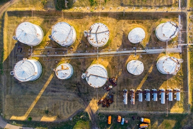 An aerial view of a large oil refinery complex with numerous storage tanks, pipelines, and industrial structures, emitting some smoke and steam into the atmosphere, all under a clear blue sky.